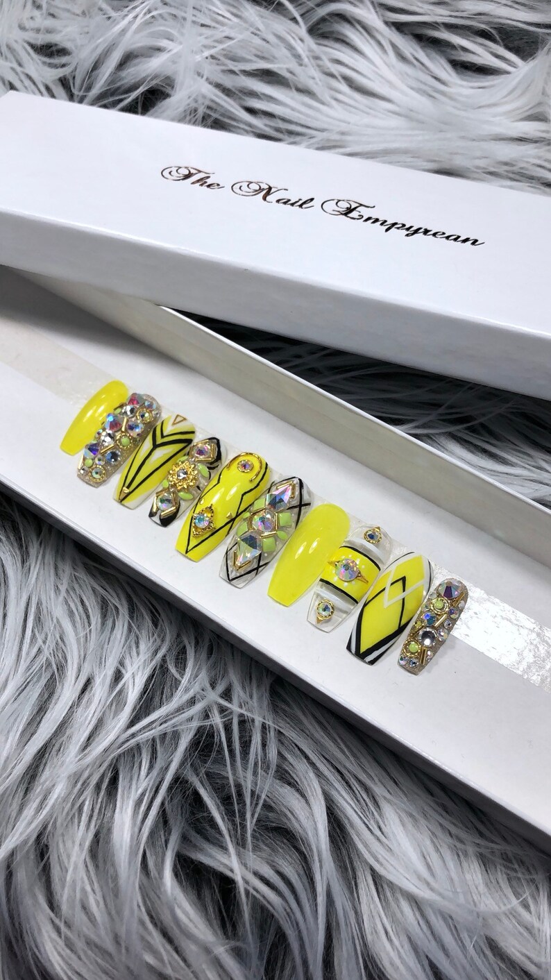 May include: A set of ten yellow press-on nails with black and white geometric designs and rhinestones. The nails are in a white box with the text "The Nail Empyrean".