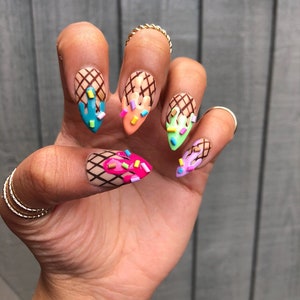 May include: Close-up of a hand with five nails painted with an ice cream cone design. Each nail features a different color ice cream with sprinkles and a waffle cone base.