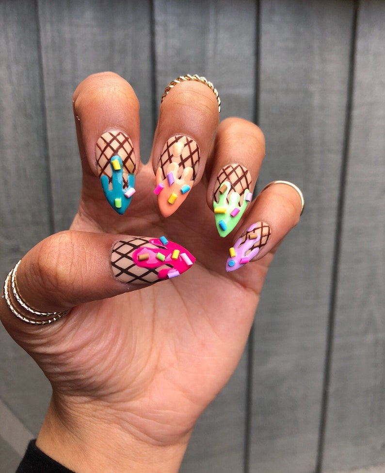 May include: Close-up of a hand with five nails painted with an ice cream cone design. Each nail features a different color ice cream with sprinkles and a waffle cone base.