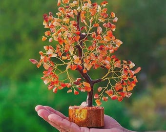May include: A decorative gemstone tree with a brown trunk and branches, adorned with orange and red gemstone leaves. The tree sits in a small wooden pot and is held in a hand against a blurred green background.