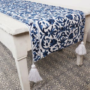 May include: A white table runner with a blue floral embroidery pattern. The runner has tassels on each end and is draped over a white table. The table is sitting on a black and white patterned rug.