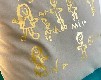 Peut inclure: Une taie d'oreiller gris clair avec des dessins d'enfants sérigraphiés en doré représentant des personnages en bâtonnets et des noms comme Ivy, Barney, Archie, Milo et Nana. Le tissu a une texture lisse et l'encre dorée crée un contraste.