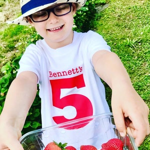 Op de afbeelding: Een wit T-shirt met rode tekst die "Bennett's" zegt en een groot rood nummer "5". Een kind draagt het shirt en houdt een bakje met aardbeien.