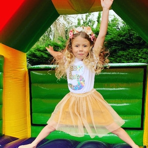 May include: A young girl wearing a white t-shirt with a glittery number "5" on it, a gold tulle skirt, and a flower crown. She is jumping in a bouncy castle with a green and yellow exterior.