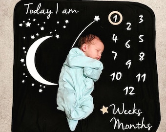 Puede incluir: Una manta negra para bebé con estrellas blancas, una luna creciente y el texto "Today I am" y "Weeks Months". Un bebé está durmiendo en la manta, y un círculo de madera con el número "1" está colocado en la manta.