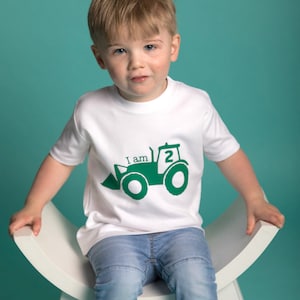 May include: A white t-shirt with a green tractor design and the text "I am 2". A young child is wearing the shirt and sitting on a white stool.