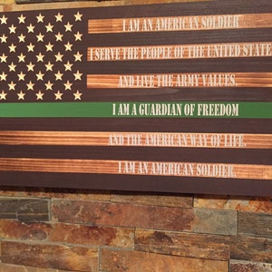 May include: A wooden American flag with a green stripe across the middle. The flag has the words "I am an American Soldier, I serve the people of the United States and live the Army values. I am a guardian of freedom and the American way of life. I am an American Soldier." written on it.