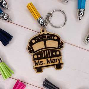 May include: A wooden school bus keychain with the text "SCHOOL BUS" and "Ms. Mary" engraved on it. The keychain has a yellow tassel and a silver keyring. Other keychains with colorful tassels are in the background.