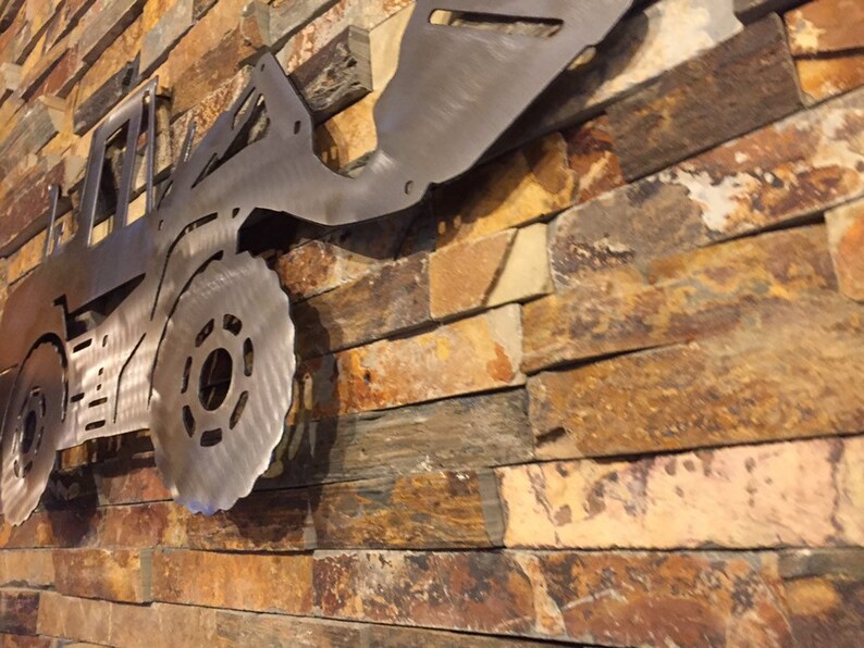May include: A metal silhouette of a tractor mounted on a stone wall. The tractor is made of brushed metal and has a detailed design.