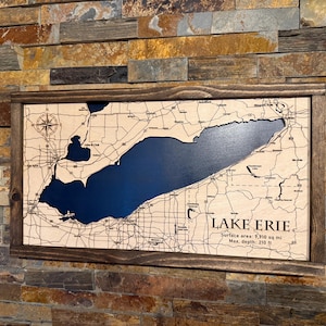 May include: A wooden map of Lake Erie, framed in a dark brown wood. The lake is a deep blue, contrasting with the light wood of the surrounding land. The map includes a compass rose and text that reads "Lake Erie".