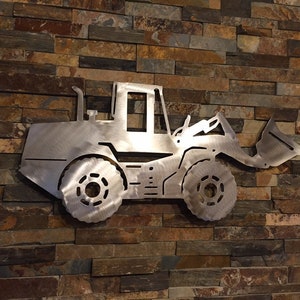 May include: A metal silhouette of a front loader excavator, mounted on a stone wall. The silhouette is cut out of a silver metal sheet.