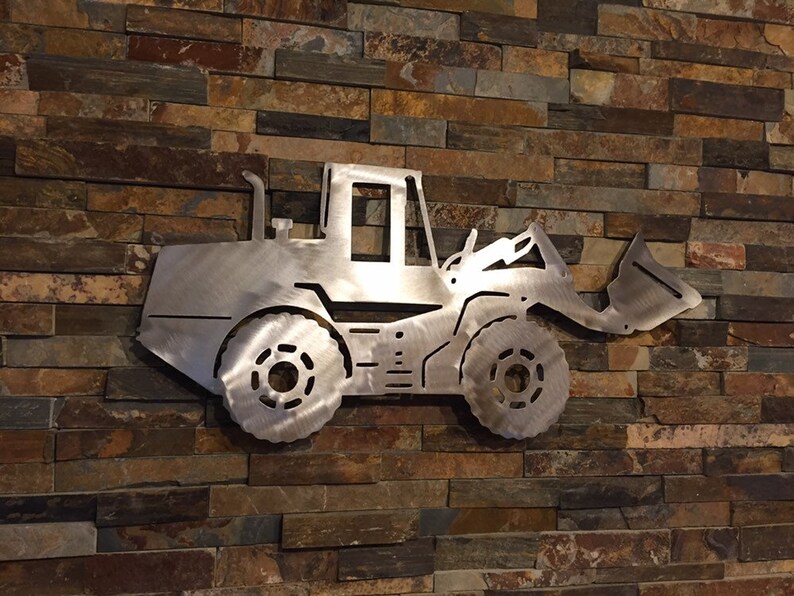 May include: A metal silhouette of a front loader excavator, mounted on a stone wall. The silhouette is cut out of a silver metal sheet.