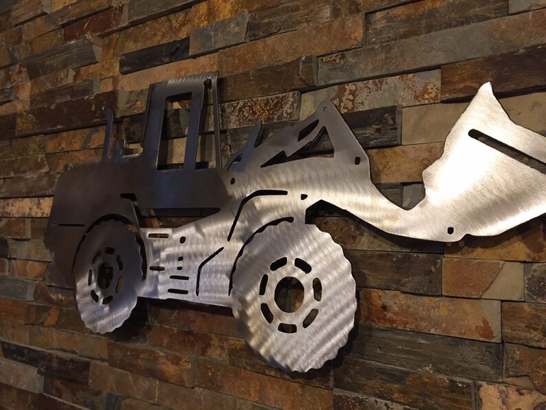 May include: A metal wall sculpture of a front loader, a type of construction equipment, with a silver finish. The sculpture is mounted on a brick wall.