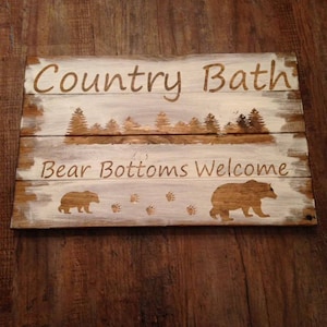 May include: A rustic wooden sign with white paint and brown lettering. The sign reads "Country Bath Bear Bottoms Welcome" with a silhouette of a bear and paw prints.