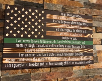 May include: A wooden sign with a distressed wood finish features a stylized American flag with a green stripe across the bottom. The sign includes a text quote about the values of an American soldier.
