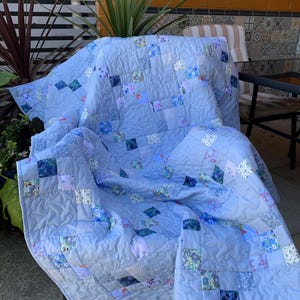 Pode incluir: Uma colcha de retalhos com tecido azul-claro s&oacute;lido intercalado com pequenos blocos quadrados em v&aacute;rios padr&otilde;es florais e geom&eacute;tricos. A colcha tem uma textura acolchoada com costuras vis&iacute;veis e est&aacute; drapeada sobre uma cadeira.