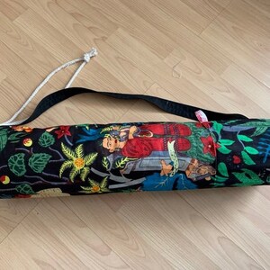 May include: A black yoga mat bag with a colorful floral and figure print. The bag has a white drawstring closure and a black adjustable strap.