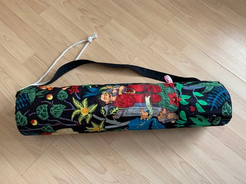 May include: A black yoga mat bag with a colorful floral and figure print. The bag has a white drawstring closure and a black adjustable strap.