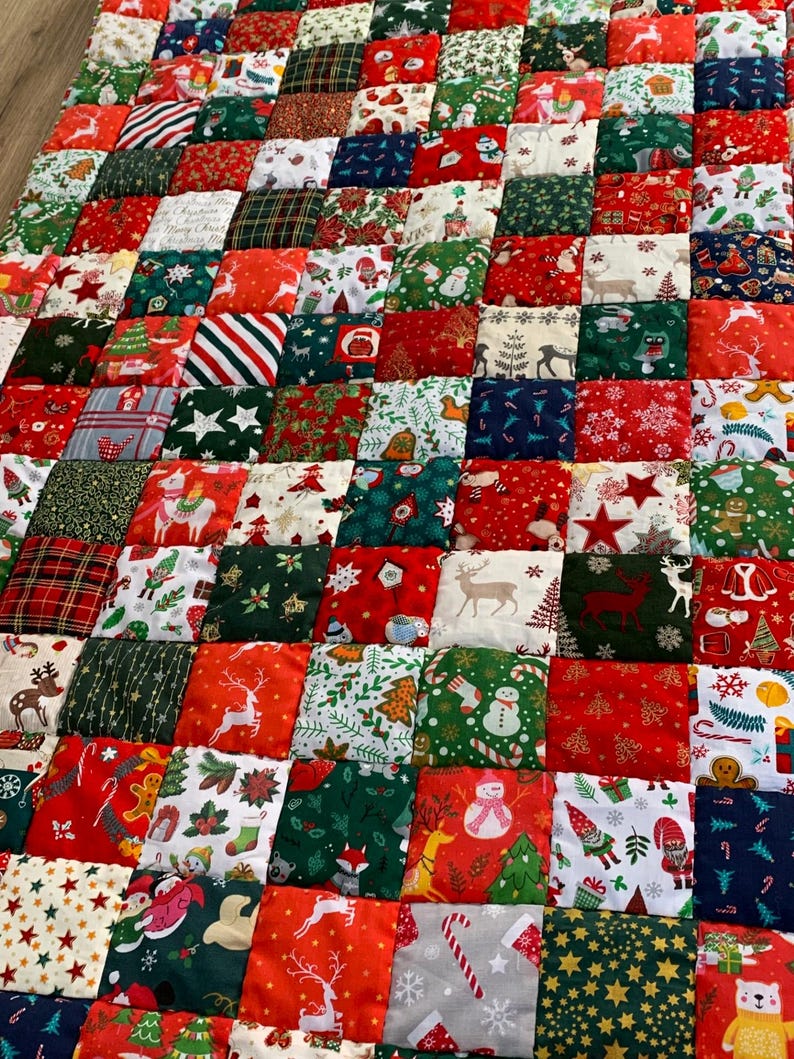 May include: A patchwork quilt with a festive Christmas theme. The quilt is composed of numerous fabric squares in red, green, white, and navy blue. Each square features holiday-themed patterns, including reindeer, snowmen, and Christmas trees.