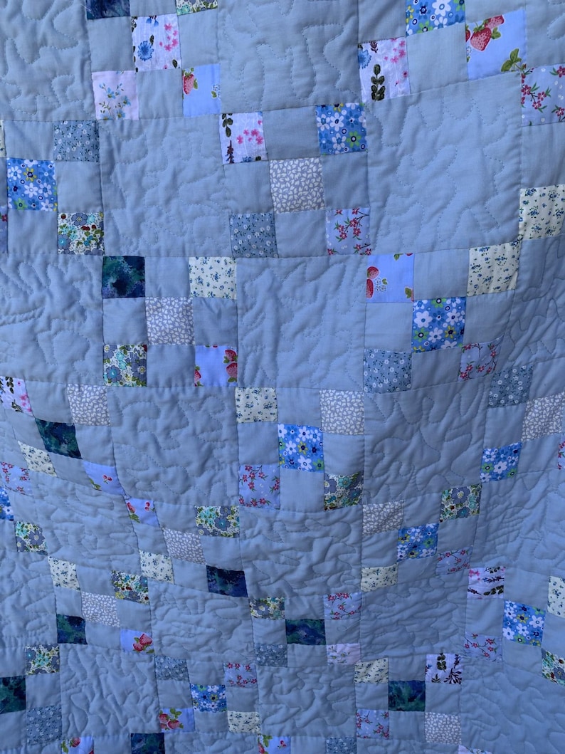 Pode incluir: Uma colcha de retalhos com um padr&atilde;o repetitivo de quadrados azul-claros s&oacute;lidos intercalados com quadrados menores de v&aacute;rias estampas florais e bot&acirc;nicas em tons de azul, branco, rosa e verde, com costuras acolchoadas onduladas.