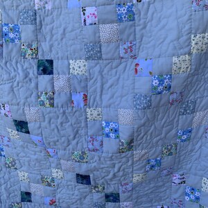 Pode incluir: Uma colcha de retalhos com um padr&atilde;o repetitivo de quadrados azul-claros s&oacute;lidos intercalados com quadrados menores de v&aacute;rias estampas florais e bot&acirc;nicas em tons de azul, branco, rosa e verde, com costuras acolchoadas onduladas.