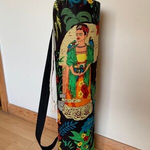 May include: Black yoga mat bag with a colorful print featuring a woman with flowers and birds. The bag has a drawstring closure and a black strap.