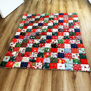 May include: A festive patchwork quilt with a Christmas theme. The quilt is made up of many small, square fabric patches in red, green, white, and blue. The patches feature holiday-themed patterns, including stars, reindeer, and Christmas trees. The quilt is on a wooden floor.
