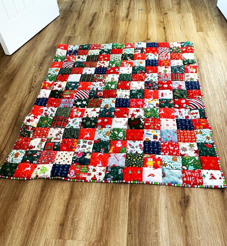 May include: A patchwork quilt with a festive Christmas theme. The quilt is composed of numerous small, square fabric patches in red, green, white, and blue. The patches feature holiday-themed patterns, including stars, reindeer, and Christmas trees. The quilt is lying on a wooden floor.
