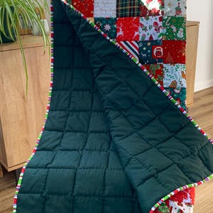 May include: A Christmas-themed quilt with a patchwork design. The top features various festive patterns in red, green, and white squares. The quilt's underside is a solid dark green, and the edges are trimmed with a striped border.