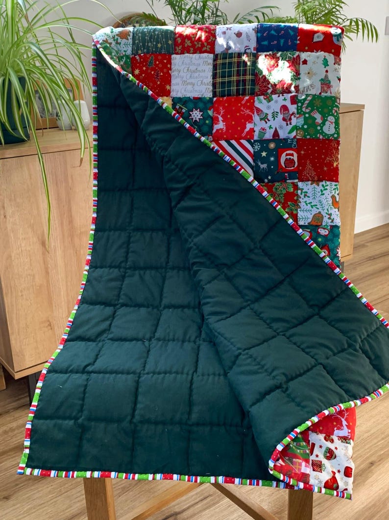 May include: A Christmas-themed quilt with a patchwork design. The top features various festive patterns in red, green, and white squares. The quilt's underside is a solid dark green, and the edges are trimmed with a striped border.