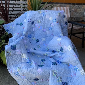 May include: A patchwork quilt featuring a light blue solid fabric interspersed with small square blocks in various floral and geometric patterns. The quilt has an overall quilted texture with visible stitching and is draped over a chair.