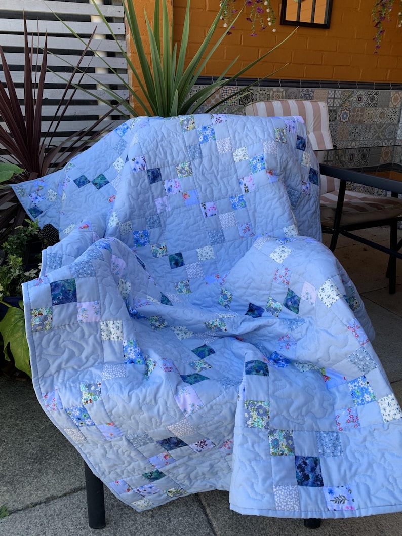 Pode incluir: Uma colcha de retalhos com tecido azul-claro s&oacute;lido intercalado com pequenos blocos quadrados em v&aacute;rios padr&otilde;es florais e geom&eacute;tricos. A colcha tem uma textura acolchoada com costuras vis&iacute;veis e est&aacute; drapeada sobre uma cadeira.