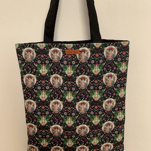 May include: Black tote bag with a floral pattern featuring a repeated image of Frida Kahlo. The bag has black straps and a brown leather tag that says "Hand Made".