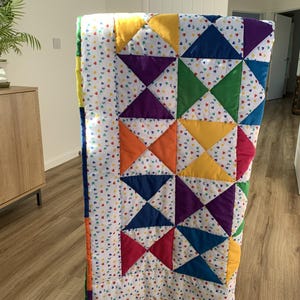 May include: A colorful quilt with a patchwork design. The quilt features a pattern of triangles in various colors, including yellow, orange, blue, green, and purple, set against a white background dotted with tiny hearts. The edges are trimmed with a rainbow border.