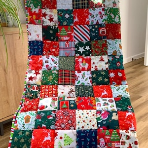 May include: A Christmas-themed patchwork quilt draped over a chair. The quilt features a variety of festive patterns, including reindeer, stars, and Christmas trees, in red, green, and white squares. The edges are trimmed with a striped border.