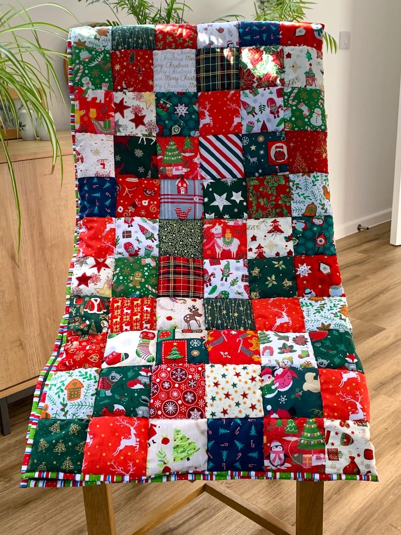May include: A Christmas-themed patchwork quilt draped over a chair. The quilt features a variety of festive patterns, including reindeer, stars, and Christmas trees, in red, green, and white squares. The edges are trimmed with a striped border.