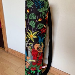 May include: Black yoga mat bag with a colorful floral and botanical print featuring a woman with a cat. The bag has a long black strap.