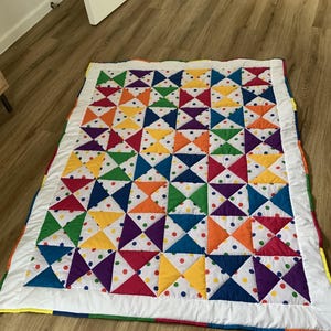 Peut inclure: Une courtepointe colorée avec un motif géométrique de triangles et de pois de différentes couleurs, dont le rouge, l'orange, le jaune, le vert, le bleu et le violet. La courtepointe a une bordure blanche et est posée sur un sol en bois.