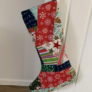 May include: A Christmas stocking constructed from various fabric patches featuring festive patterns including red snowflakes, green holly, striped ribbons, and stars. The stocking is suspended by a thin red cord and displays a colourful patchwork design.