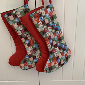 May include: Two Christmas stockings featuring a patchwork pattern of festive icons including Santa Claus, reindeer, snowmen, and plaid squares. The stockings have solid red fabric backs and red ribbon loops for hanging, with a multi-colored front design.