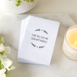 May include: A stack of white cards with black text that reads "Plant these seeds and grow happy memories". The text is surrounded by a wreath of leaves and a small heart.