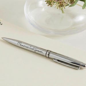 May include: A silver pen with the inscription "Memories last forever." The pen is lying on a cream-colored surface next to a spiral-bound notebook and a glass vase with flowers.