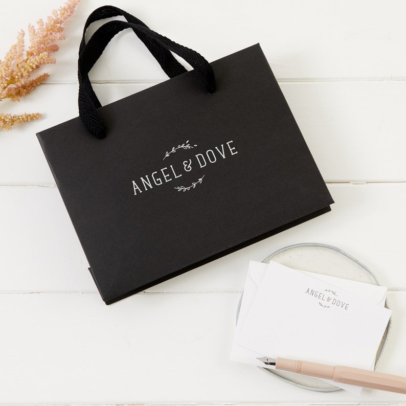 Puede incluir: Una bolsa de regalo negra con un logotipo blanco que dice "Angel & Dove" y una etiqueta de regalo blanca con el mismo logotipo.