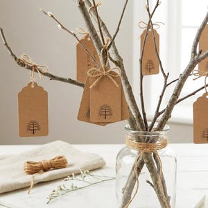 May include: A clear glass vase holds tree branches adorned with brown paper tags. Each tag features a tree illustration and is tied with twine. A roll of twine and a linen cloth are visible.