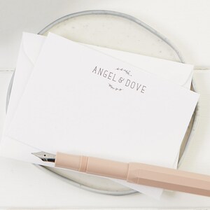 Puede incluir: Una tarjeta blanca con el texto "ANGEL & DOVE" en una fuente de letra manuscrita simple. El texto est&aacute; rodeado por una corona de peque&ntilde;as hojas. Un bol&iacute;grafo rosa est&aacute; sobre la tarjeta.