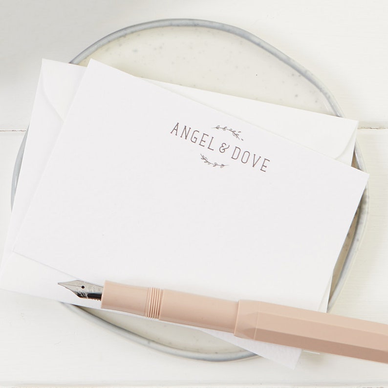 Puede incluir: Una tarjeta blanca con el texto "ANGEL & DOVE" en una fuente de letra manuscrita simple. El texto est&aacute; rodeado por una corona de peque&ntilde;as hojas. Un bol&iacute;grafo rosa est&aacute; sobre la tarjeta.