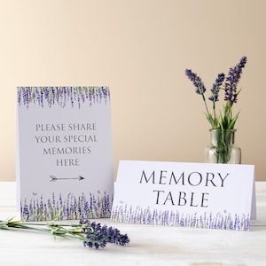 May include: White card signs with lavender floral borders. One sign reads "PLEASE SHARE YOUR SPECIAL MEMORIES HERE" with an arrow. The other says "MEMORY TABLE". A vase of lavender and sprigs of lavender are also visible.