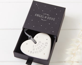 May include: A white ceramic heart ornament with the embossed text LOVED MISSED REMEMBERED, hanging from a black and white striped ribbon. The heart is presented inside a black sliding gift box featuring the text ANGEL & DOVE and small stars.