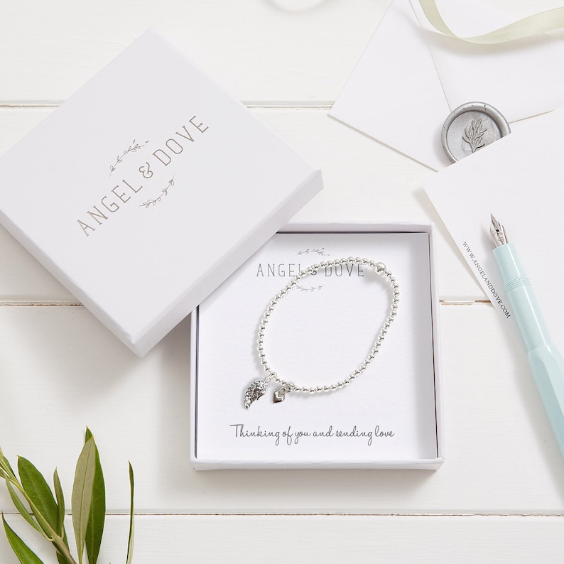 Puede incluir: Una caja de regalo blanca con un brazalete de plata en su interior. El brazalete tiene un peque&ntilde;o dije de plata en forma de paloma y las palabras "Thinking of you and sending love" est&aacute;n impresas en la caja.