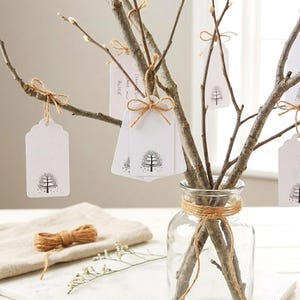 May include: A clear glass jar holds tree branches with white tags featuring a black tree design. The tags are tied to the branches with twine bows. A roll of twine and a linen cloth are in the foreground, suggesting a rustic, natural theme.